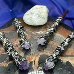 May include: Four handcrafted wands with silver-toned metalwork and amethyst crystal tips. Each wand has a different colored handle: green, blue, brown, and purple. A white stone sits in the background on a dark, patterned surface.