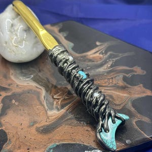 May include: A handcrafted crochet hook with a gold-colored hook and a dark metal handle. The handle is adorned with turquoise and white stones, and a decorative design. The hook rests on a decorative surface.