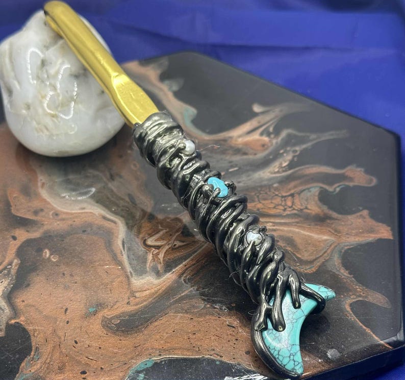May include: A handcrafted crochet hook with a gold-colored hook and a dark metal handle. The handle is adorned with turquoise and white stones, and a decorative design. The hook rests on a decorative surface.