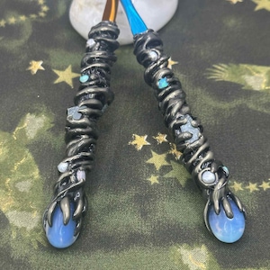 May include: Two handcrafted wands with dark gray, textured handles adorned with small, iridescent gems. The wands have blue and brown shafts and end in a blue, teardrop-shaped crystal. The wands are set against a green background with gold stars.