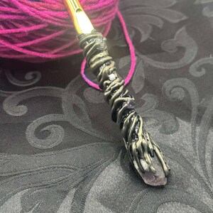 May include: A handcrafted crochet hook with a gold-colored hook and a decorative handle. The handle is wrapped with bronze-colored metal and features a purple crystal at the base. A ball of colorful yarn is in the background.