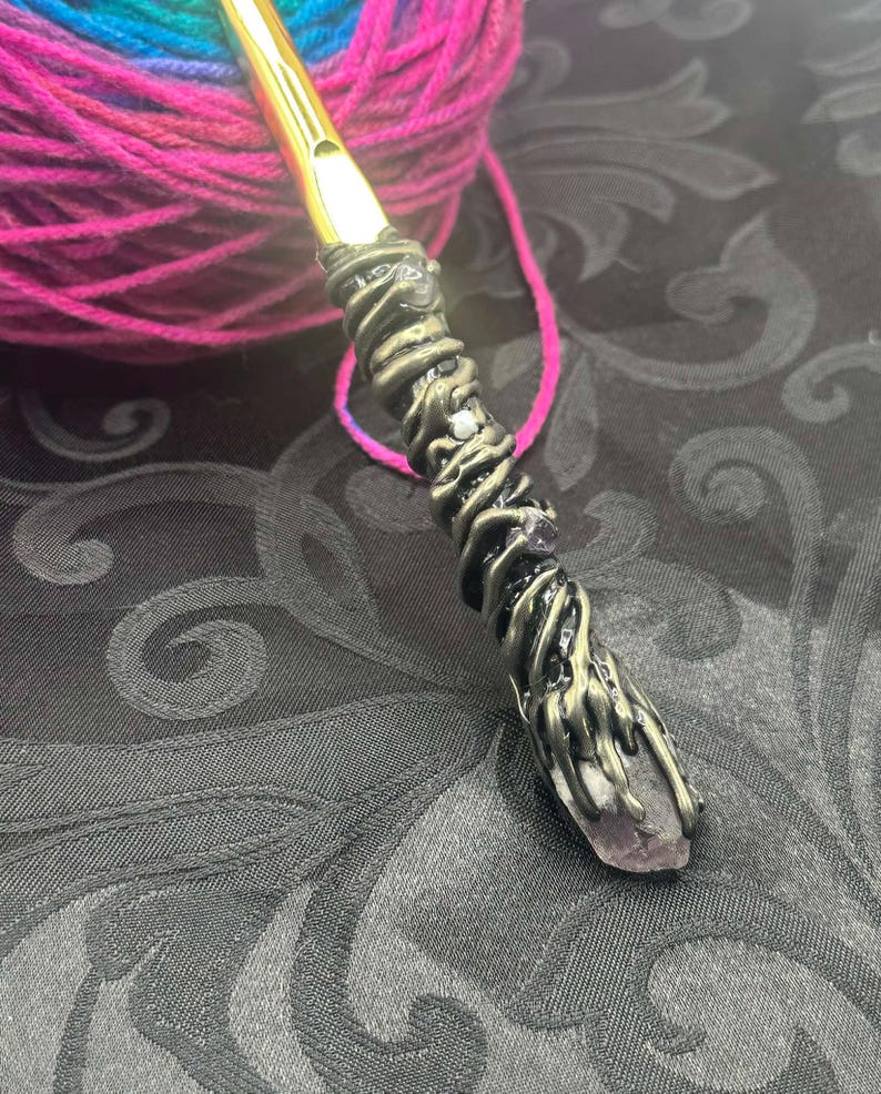 May include: A handcrafted crochet hook with a gold-colored hook and a decorative handle. The handle is wrapped with bronze-colored metal and features a purple crystal at the base. A ball of colorful yarn is in the background.