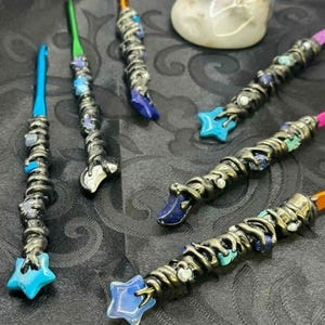 May include: A collection of handcrafted crochet hooks with decorative metalwork and colorful accents. Each hook features a unique design, including star-shaped and gemstone embellishments. The hooks have various colored tips and are arranged on a dark surface.