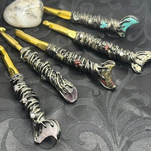 May include: A collection of handcrafted crochet hooks with ornate metal handles and gold-colored hook ends. The handles are embellished with various gemstones and decorative elements, including turquoise, amethyst, and pearls. The hooks are arranged on a dark, patterned surface.