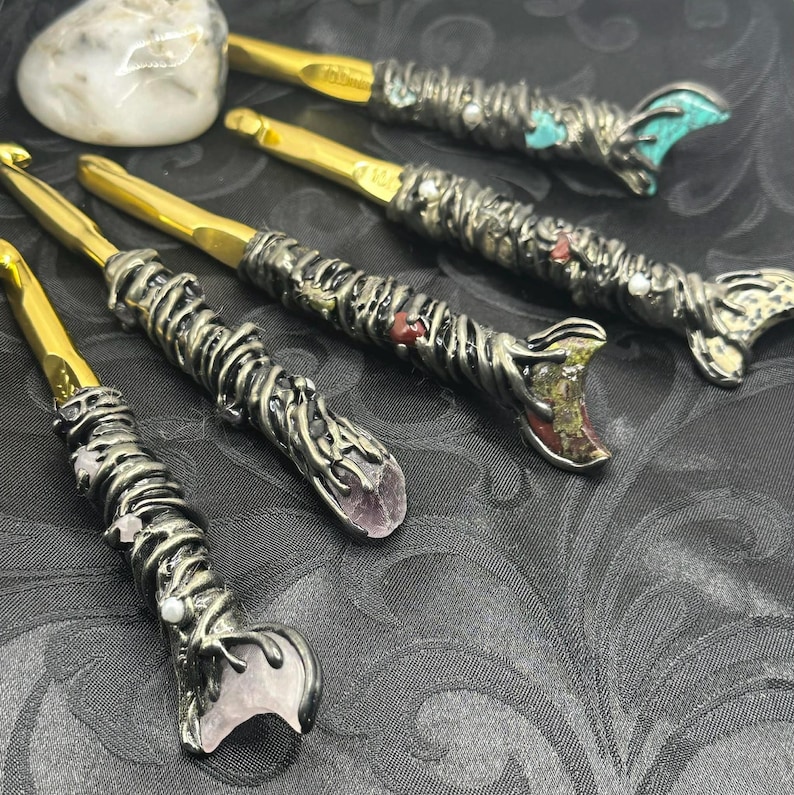 May include: A collection of handcrafted crochet hooks with ornate metal handles and gold-colored hook ends. The handles are embellished with various gemstones and decorative elements, including turquoise, amethyst, and pearls. The hooks are arranged on a dark, patterned surface.