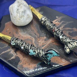 May include: Two crochet hooks with gold-colored tips and ornate, silver-toned handles. The handles are embellished with turquoise stones, white beads, and a textured design. A white stone rests nearby on a decorative surface.