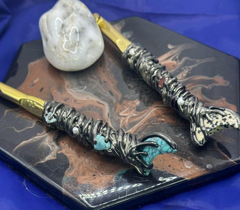 May include: Two crochet hooks with gold-colored tips and ornate, silver-toned handles. The handles are embellished with turquoise stones, white beads, and a textured design. A white stone rests nearby on a decorative surface.
