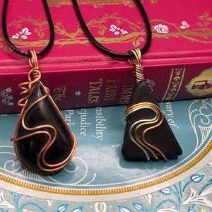 May include: Two necklaces with black stone pendants wrapped in copper wire. One pendant is a teardrop shape and the other is a triangular shape. The necklaces are displayed on a book with the title "Emma's Fairy Tales".