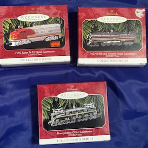 May include: Three Hallmark Keepsake ornaments featuring miniature train locomotives. The ornaments are in red boxes with green sprigs. The top left box shows a red and silver 1950 Santa Fe F3 Diesel Locomotive. The top right box shows a black and red 746 Norfolk and Western Steam Locomotive. The bottom box shows a Pennsylvania GG-1 Locomotive.