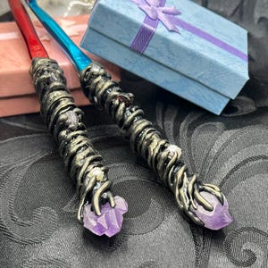 May include: Two crochet hooks with metallic red and blue shafts, featuring custom handles wrapped in dark, textured, vine-like material. Each handle is accented with small embedded stones and topped with a raw, light purple amethyst crystal point.