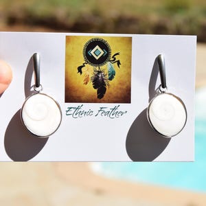Può includere: Un paio di orecchini color argento con un design bianco a spirale all'interno di una cornice circolare. Gli orecchini pendono da un anello d'argento. L'immagine mostra anche una carta con una grafica acchiappasogni e il testo "Ethnic Feather".