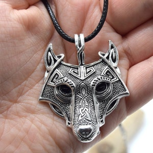 Könnte beinhalten: Ein silberner Wolfskopf-Anhänger mit filigranen Details und einer schwarzen Lederschnur. Der Anhänger zeigt ein keltisches Knotendesign und ein Wolfsgesicht mit detaillierter Schnauze und Augen.