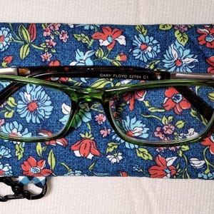 May include: A blue and white checked fabric eyeglass case with a floral print on the front. The case has a metal clasp and a pair of green eyeglasses with black frames are resting on top of the case. The eyeglasses have the text "GARY FLOYD 52754 C1" printed on the frame.