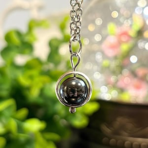 May include: A silver-toned necklace featuring a dark, spherical pendant encased in a circular frame. The pendant has a reflective, metallic appearance. The chain is delicate, and the background is blurred with green foliage and soft lighting.