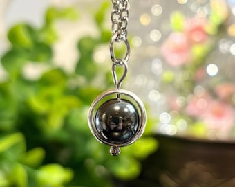 Puede incluir: Un collar plateado con un colgante esférico oscuro, encerrado en un marco circular. El colgante tiene una apariencia metálica reflectante. La cadena es delicada, y el fondo está borroso con follaje verde e iluminación suave.