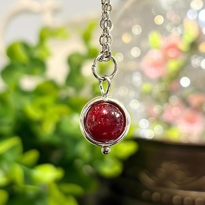 May include: A silver pendant necklace featuring a round, deep red gemstone. The gemstone is set in a silver bezel and hangs from a delicate silver chain. The background is blurred, with green foliage and soft pink flowers.