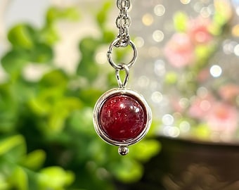 Puede incluir: Un collar con colgante de plata con una piedra preciosa redonda de color rojo intenso. La piedra preciosa está engastada en un bisel plateado y cuelga de una delicada cadena plateada. El fondo está borroso, con follaje verde y flores rosas suaves.