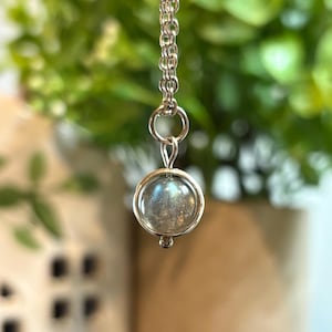 May include: A silver necklace featuring a round, iridescent pendant. The pendant is encased in a silver frame and hangs from a delicate chain. The background is blurred, with green foliage and a neutral-toned surface.