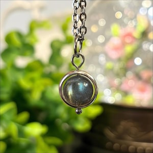 May include: A silver-toned metal necklace featuring a circular pendant frame that holds a round, polished labradorite gemstone. The stone displays a dark grey base color with a distinct iridescent blue flash. The pendant hangs from a simple link chain.