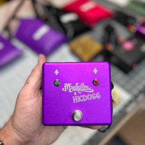 May include: A purple Madhaller x HICDOGG effects pedal. The square pedal has a silver button, two indicator lights, and white text. The top of the pedal has up and down arrows. The pedal is held in a hand.