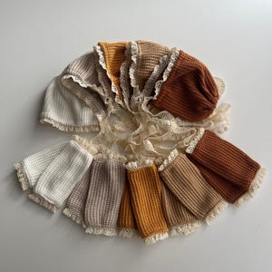 May include: A collection of baby bonnets and matching arm warmers in various neutral colors, including white, beige, mustard, and rust. Each bonnet and arm warmer features a waffle knit texture and is trimmed with delicate lace.