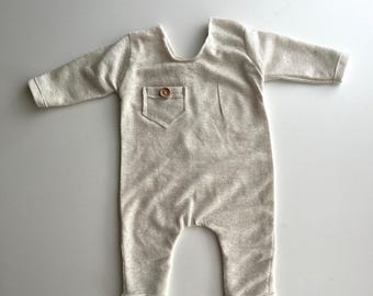 May include: A light beige baby romper with long sleeves and a small pocket on the chest. The romper has a round wooden button on the pocket and is made of a soft, textured fabric. The legs are cuffed at the ankles.