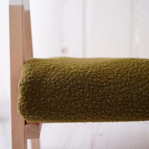 Può includere: Primo piano di una sedia in legno con una seduta in tessuto verde. Il tessuto ha una superficie testurizzata e pelosa.