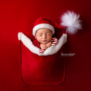 May include: A newborn baby wearing a red Santa hat and a white fur-trimmed outfit is sleeping in a red Santa sack. The baby is holding a candy cane in their hands.