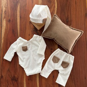 May include: A newborn photography set featuring a white textured onesie and pants with brown pockets, a matching hat, and a brown pillow. The set is designed for baby photoshoots.