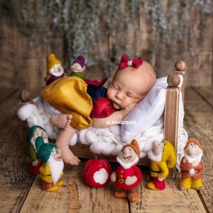 Puede incluir: Un bebé recién nacido, vestido como Blancanieves, durmiendo en una cama de madera en miniatura con una manta y almohada blancas. El bebé está rodeado de figuras de enanos de fieltro y una manzana roja. Un lazo rojo adorna la cabeza del bebé. La palabra "GLAMORGEN" es visible.