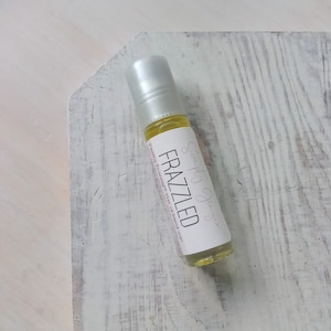 May include: A small, clear glass bottle with a silver roller ball top. The bottle is labelled "Simply Ginger" and "Frazzled" with the text "Frazzled" in bold. The bottle is sitting on a white wooden surface.