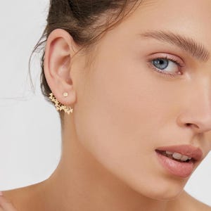 Puede incluir: Primer plano de una oreja adornada con un pendiente dorado. El pendiente presenta un pequeño perno y un diseño decorativo en forma de abanico con múltiples elementos triangulares. El tono de piel es claro y el fondo es de color neutro.