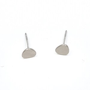 Könnte beinhalten: Ein Paar kleine, silberfarbene Ohrstecker. Jeder Ohrring hat ein glattes, kieselsteinförmiges Design. Die Ohrringe sind vor einem hellweißen Hintergrund platziert, was ihre minimalistische Ästhetik hervorhebt.