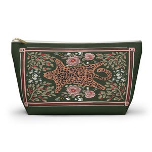 May include: A dark green zippered pouch with a decorative design. The design features a leopard print animal, pink roses, and floral elements within a rectangular frame. The zipper is gold-coloured.