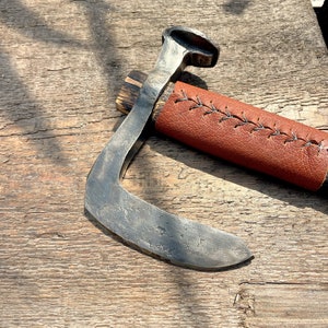 May include: A hand-forged, curved axe with a brown wooden handle and a brown leather sheath with stitching. The axe is resting on a wooden surface.