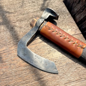 May include: A hand-forged axe with a curved blade and a brown leather handle with stitching. The axe is resting on a wooden surface.