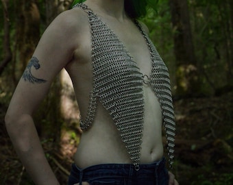 May include: A woman with teal hair wearing a silver chainmail top and blue jeans. She is standing in a forest.
