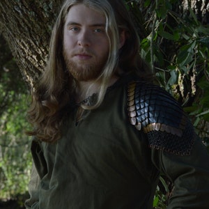 May include: A man with long blonde hair wearing a dark green shirt with a leather and metal shoulder piece. He is standing in front of a tree.