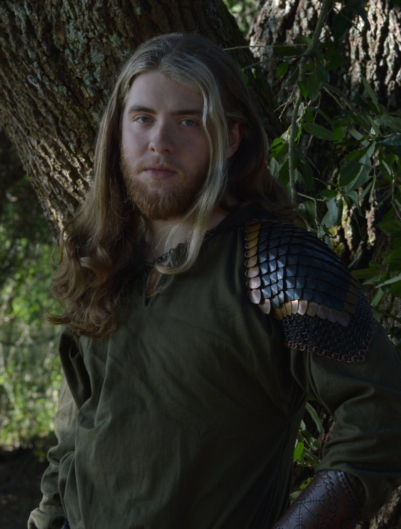 May include: A man with long blonde hair wearing a dark green shirt with a leather and metal shoulder piece. He is standing in front of a tree.