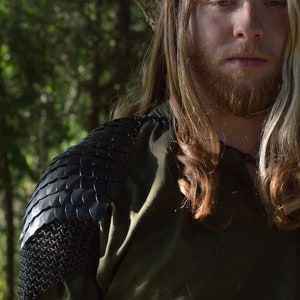 May include: A man with long blonde hair and a beard is wearing a green shirt with a black chainmail shoulder piece. He is standing in front of a tree.