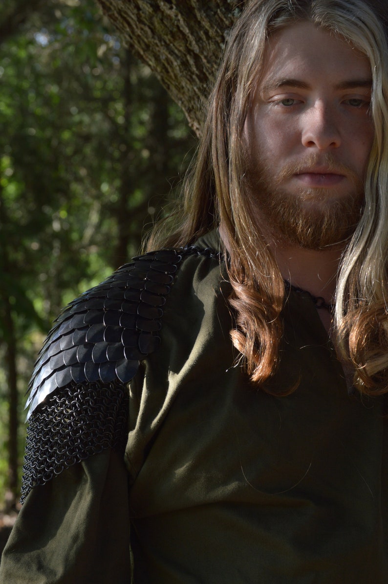 May include: A man with long blonde hair and a beard is wearing a green shirt with a black chainmail shoulder piece. He is standing in front of a tree.