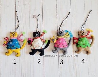 May include: Four handmade cat ornaments with black string hangers. Each cat is dressed in a sweater and scarf, and holds a small item. The cats are yellow, black, gray, and brown. The text "ARIBELLA COLLECTION" is visible.