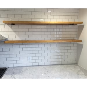 May include: Two wooden floating shelves mounted on a white subway tile backsplash. The shelves are a light brown color and appear to be made of solid wood. The kitchen counter is visible in the foreground.