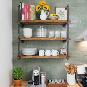 May include: A three-tier wall-mounted shelving unit made of dark metal pipes and wooden planks. The shelves hold books, a white pitcher with yellow sunflowers, a glass jar, clear glasses, white bowls, white mugs, and a framed botanical print.