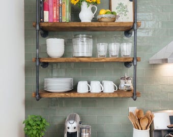 May include: A three-tier wall-mounted shelving unit made of dark metal pipes and wooden planks. The shelves hold books, a white pitcher with yellow sunflowers, a glass jar, clear glasses, white bowls, white mugs, and a framed botanical print.