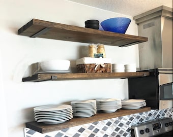 May include: Three horizontal dark brown wooden shelves mounted on a wall. The top shelf holds two black bowls and a blue glass bowl. The middle shelf features a white bowl, a woven basket, and two small white ramekins. The bottom shelf holds stacks of white plates.