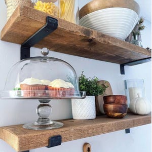 May include: Wooden shelves with black metal brackets hold various kitchen items. A glass cake stand with cupcakes is on the bottom shelf. Other items include bowls, a white pumpkin, and a small potted plant. The shelves are made of rustic, stained wood.