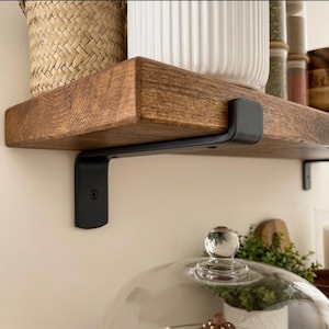 May include: A thick, medium-brown wooden floating shelf is mounted to a wall with a matte black metal bracket. The shelf features a visible wood grain texture and is secured with a visible screw. The bracket has a smooth, curved design.