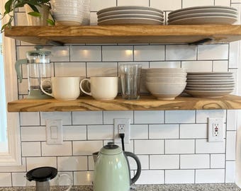 May include: Two natural wood floating shelves mounted on a white subway tile wall. The top shelf holds stacks of white ceramic bowls and plates. The bottom shelf features a sage green French press, two cream mugs, a glass, and more ceramic bowls.