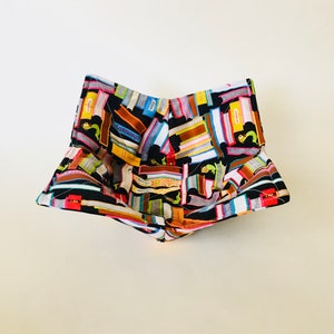 May include: A fabric bowl with a colorful book print. The bowl is black with a pattern of stacked books in various colors, including red, blue, yellow, and brown. The bowl is square-shaped with a folded design.